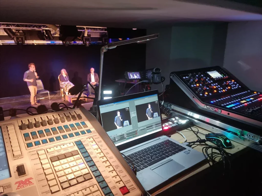 Regard sur le poste de contrôle technique avec ordinateurs et consoles de mixage éclairées lors d'un événement en direct, illustrant les services de production vidéo de MediaSmart.