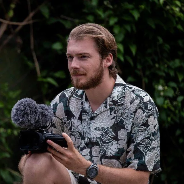 Journaliste indépendant nommé Raphael tenant un micro et une caméra, avec une expression détendue, portant une chemise à motifs dans un cadre extérieur verdoyant.